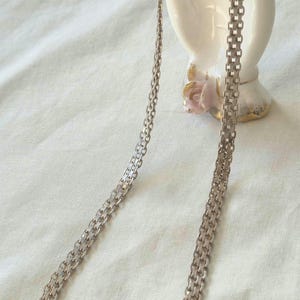 May include: A silver-toned metal chain necklace featuring a repeating interlocking link pattern. The necklace is draped in a long, continuous loop, showcasing a metallic finish and a circular spring ring clasp at the top of the chain structure.