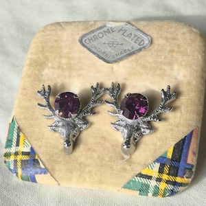 May include: A pair of chrome-plated stag head earrings with purple gemstone accents, presented on a beige velvet display. The earrings are set against a backdrop with a decorative label that reads "Chrome Plated Untarnishable".