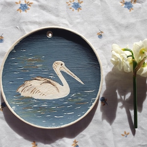 May include: A round ceramic wall plaque featuring a hand-painted white pelican swimming on dark blue water. The plaque has a small hole at the top for hanging. The pelican is depicted in profile with textured brushstrokes, and the surface of the plaque shows visible paint application marks throughout the blue water area.
