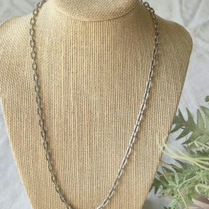 May include: A silver-toned metal chain necklace featuring elongated, rectangular links with rounded edges. The necklace is displayed on a textured, neutral-coloured bust form, showcasing the uniform, repeating pattern of the metallic chain links.