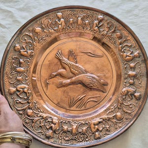 May include: A round copper plate featuring a central embossed design of two flying ducks over reeds. The wide border is decorated with intricate relief carvings of small figures, scrolling foliage, and oval accents in a warm metallic finish.