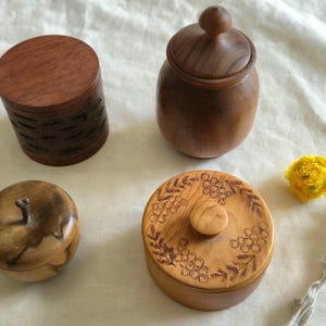 May include: Four wooden containers of different shapes and sizes. One is cylindrical with carved details, another is a small jar with a lid, a third is apple-shaped, and the last is round with a floral design on the lid. All are made of wood.