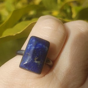 May include: A ring featuring a rectangular, deep blue lapis lazuli gemstone with visible gold-coloured flecks. The stone is set in a dark, oxidised metal bezel and mounted on a simple, matching dark metal band that encircles the finger.