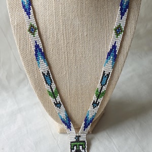 May include: A beaded necklace with a geometric pattern in white, blue, green and black. The necklace has a pendant with a stylised figure and a fringe of beads.