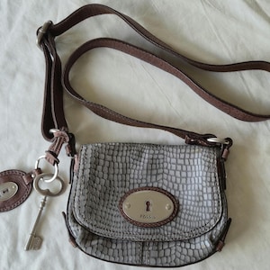 May include: A gray Fossil crossbody bag with a textured pattern, brown trim, and a long brown strap. It features a key and a leather tag with a keyhole design. The bag has a keyhole emblem on the front.