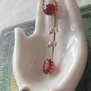 May include: A gold pendant necklace featuring three red gemstones and leaf details. The necklace is presented on a white ceramic hand. The gemstones are oval and round, and the chain is fine.