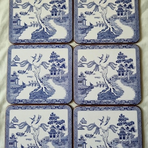 May include: A set of six square coasters, each with a blue and white Willow pattern design. The coasters depict a detailed landscape scene with pagodas, trees, and a winding path. They have a brown border.
