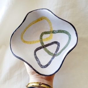 May include: A white ceramic bowl with a wavy edge and an abstract design of overlapping yellow, green, and black shapes.