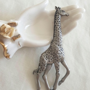 May include: A silver-toned metal giraffe brooch featuring a detailed textured coat with dark recessed spots. The giraffe is depicted in a walking pose with its head held high and neck extended, showcasing a realistic silhouette and metallic finish.