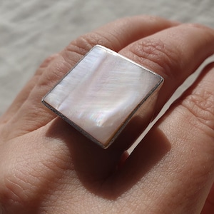 May include: A square ring featuring a mother-of-pearl inlay. The ring has a silver-coloured band and a large, diamond-shaped face. The iridescent surface of the inlay reflects light, creating a shimmering effect. The ring is worn on a finger.
