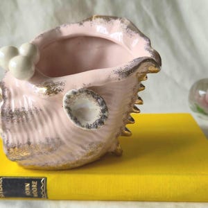 May include: A pink ceramic seashell-shaped planter with gold trim and a cluster of white spheres. The planter sits on a yellow book with the title "Dance and Skylark" by John Moore. A glass paperweight with a pink flower is in the background.