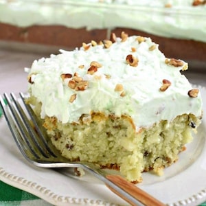 May include: A slice of green coconut cake with white icing and chopped pecans on a white plate with a fork.