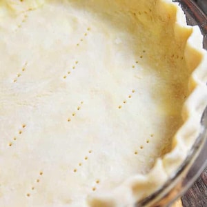 May include: A close-up shot of a pie crust in a clear glass pie dish. The crust is a light golden color, with a crimped edge and small holes. The pie crust is ready for filling and baking.