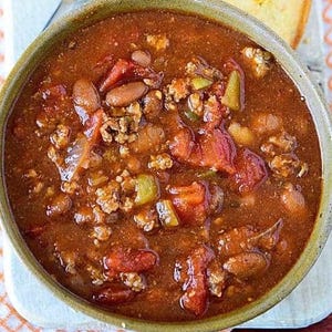 Könnte beinhalten: Nahaufnahme einer Schüssel mit Chili. Das Chili hat eine tiefrote bis braune Farbe und enthält Tomatenstücke, Bohnen und Hackfleisch. Ein Stück Maisbrot ist an der Seite der Schüssel zu sehen.