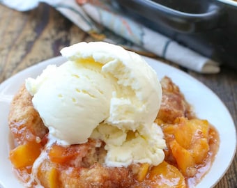 May include: A portion of peach cobbler with a scoop of vanilla ice cream on a white plate. The cobbler features a golden-brown crust and is filled with peach slices in a sweet sauce. The dessert is presented on a wooden surface.