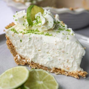 May include: A slice of key lime pie with a graham cracker crust, topped with whipped cream, lime zest, and a lime slice. The pie is on a gray plate with lime slices in the foreground. A dessert.