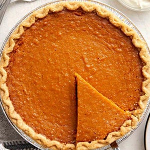May include: A whole pumpkin pie with a golden-brown crust and filling, with a slice removed. The pie is in a silver tin, and a pie server is inserted into the pie. A small bowl of whipped cream is in the background.