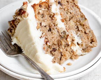 May include: A slice of banana cake with white frosting and pecans on a white plate.