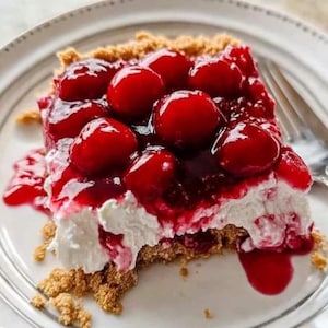 May include: A slice of cherry cheesecake on a white plate. The dessert features a graham cracker crust, a creamy white filling, and a topping of bright red cherries in a glossy sauce. A fork is visible in the background.