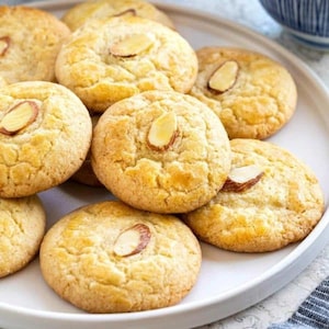Puede incluir: Un plato de galletas de almendras, de color marrón claro y ligeramente dorado. Las galletas están dispuestas en forma circular, con la almendra en la parte superior de cada galleta.