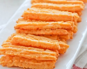 May include: A white rectangular platter piled high with orange cheese straws. The straws are arranged in a staggered pattern, with the tips of the straws pointing towards the right side of the image.