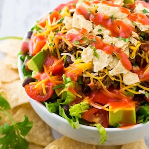 Puede incluir: Una vibrante ensalada de tacos en un cuenco blanco, con capas de lechuga fresca, tomates cortados en dados, aguacate, frijoles negros y queso rallado. La ensalada está cubierta con totopos y un chorrito de salsa roja.