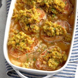May include: A white rectangular baking dish filled with cooked pork chops topped with a yellow-green stuffing. The pork chops are in a light brown gravy, with visible herbs. The dish is set on a blue and white striped cloth.
