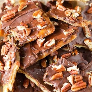 May include: A close-up of a pile of chocolate toffee bark with pecans. The bark is broken into irregular pieces and is covered in a layer of dark chocolate. The pecans are scattered throughout the bark and are visible on the surface.