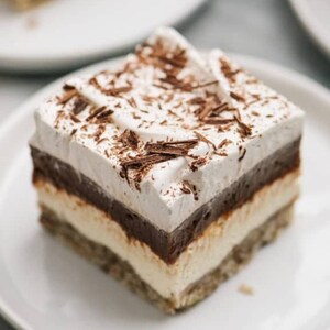 Op de afbeelding: Een vierkant stuk gelaagd dessert op een wit bord. Het dessert heeft een kruimelige basis, een romige witte laag, een donkere chocoladelaag en een topping van slagroom met chocoladeschaafsel. Een rijke, decadente traktatie.