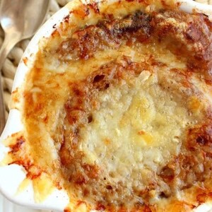 以下が含まれることがあります： フランス風オニオンスープが入った白いセラミックボウルをクローズアップした写真。スープには溶けたチーズとトーストしたパンがトッピングされています。