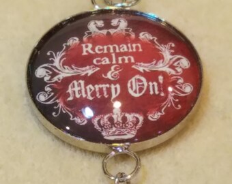 May include: A silver-toned pendant with a round, red and black design that reads "Remain calm & Merry On!" in white script. A white bead with red polka dots hangs below. The pendant has a silver loop for a chain.