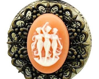 May include: A gold tone pocket watch necklace with a cameo featuring three women in white on an orange background. The watch has a detailed ornate design.