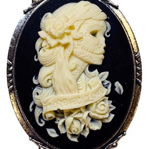 May include: A black cameo brooch with a white cameo of a woman with a skull face and flowers. The cameo has the text "Forever Love" on it.