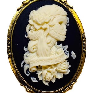 May include: A gold-toned cameo brooch with a black background and a white cameo of a woman with a skull-like face. The woman is wearing a floral crown and has long hair. The cameo is surrounded by a gold-toned frame.