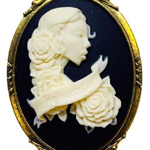May include: A gold-tone cameo brooch with a white cameo of a woman's profile with roses and a ribbon. The cameo is set against a black background.