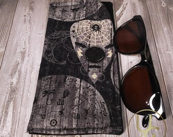 以下が含まれることがあります： 黒と白のパターンが施された布製の眼鏡ケース。黒と白のウィジャ盤のデザインと、中央にクモの巣とクモが描かれています。ケースは木製の表面に置かれ、黒いサングラスの隣にあります。