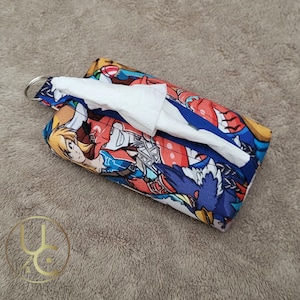 May include: A rectangular tissue holder with a colorful anime-style print featuring characters in red, blue, and yellow. The holder has a silver key ring and white tissues are visible. The holder is on a brown surface.