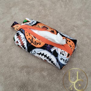 May include: A rectangular tissue holder with a Halloween-themed pattern. The fabric features orange pumpkins and white skull designs on a black background. The holder has an orange zipper and a small fabric loop.