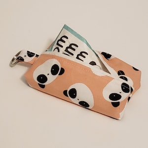 May include: A small, peach-colored fabric pouch with a panda bear pattern. The pouch has a silver key ring attached and contains a folded paper item with text. The panda bears have black eyes and ears. The pouch is designed to hold small items.