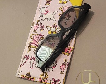 May include: Black-framed glasses rest on a pink glasses case. The case features a repeating pattern of a white cat, crowns, chairs, and perfume bottles. The case has a yellow top and is on a gray surface.