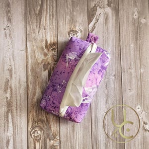 May include: Purple and white tissue box cover with a floral and butterfly design. The cover is made of fabric and has a flap to pull out tissues.