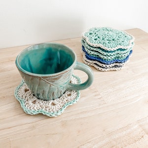 May include: A teal ceramic mug sits on a crocheted coaster with a light green and white design. A stack of crocheted coasters in various shades of green and blue are in the background.