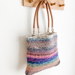 Pode incluir: Uma bolsa tote de croch&ecirc; listrada multicolorida com al&ccedil;as de couro marrom penduradas em um rack de madeira.