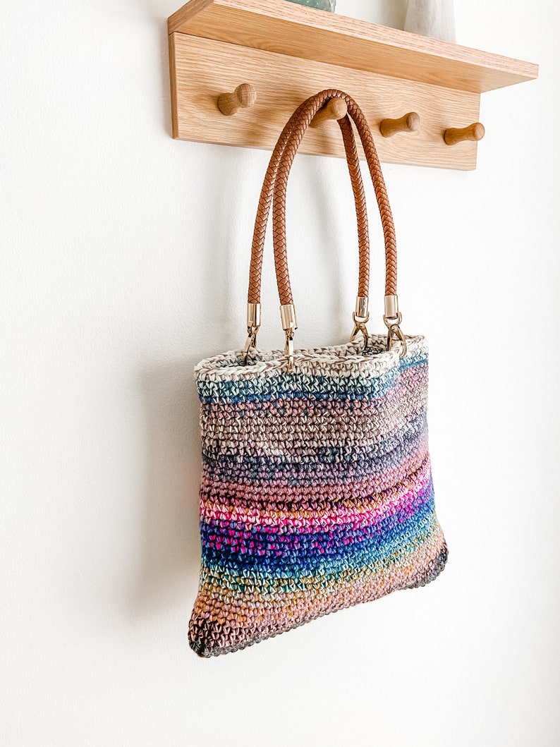 Pode incluir: Uma bolsa tote de croch&ecirc; listrada multicolorida com al&ccedil;as de couro marrom penduradas em um rack de madeira.