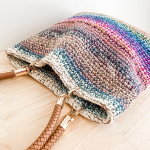 Pode incluir: Uma bolsa tote de croch&ecirc; com listras arco-&iacute;ris e al&ccedil;as de couro marrom. A bolsa tem fechos de metal dourado nas al&ccedil;as.