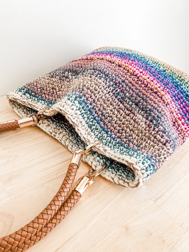 Pode incluir: Uma bolsa tote de croch&ecirc; com listras arco-&iacute;ris e al&ccedil;as de couro marrom. A bolsa tem fechos de metal dourado nas al&ccedil;as.