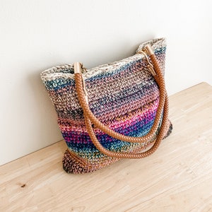 Pode incluir: Uma bolsa tote de croch&ecirc; listrada com al&ccedil;as de couro marrom e detalhes dourados. A bolsa &eacute; feita de v&aacute;rias cores, incluindo rosa, azul, verde e marrom.
