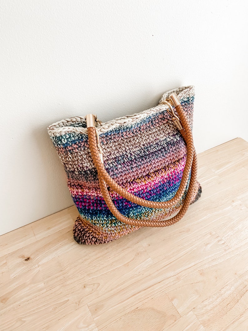 Pode incluir: Uma bolsa tote de croch&ecirc; listrada com al&ccedil;as de couro marrom e detalhes dourados. A bolsa &eacute; feita de v&aacute;rias cores, incluindo rosa, azul, verde e marrom.