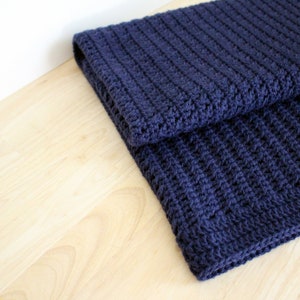 Könnte beinhalten: Eine dunkelblaue gehäkelte Decke, die in der Hälfte gefaltet ist. Die Decke ist mit einem strukturierten Stich gearbeitet und hat ein weiches, gemütliches Aussehen.
