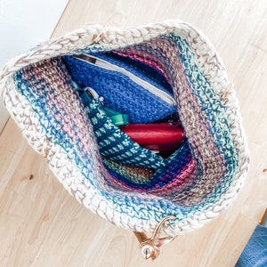 Pode incluir: Uma bolsa de ombro de croch&ecirc; com um padr&atilde;o listrado em tons de rosa, azul e verde. A bolsa tem al&ccedil;as douradas e est&aacute; cheia de v&aacute;rios itens, incluindo uma bolsa azul, uma bolsa vermelha e uma bolsa verde.